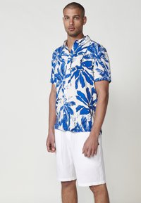 Cămașă cu mânecă scurtă, cu un model floral alb și albastru. Fabricată dintr-un material ușor. Asortată cu pantaloni scurți albi, evidențiind o croială confortabilă.