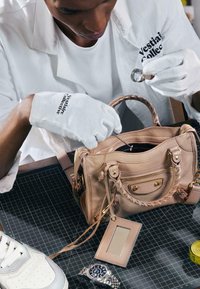Personne portant des gants blancs inspectant un sac à main beige à l'aide d'une loupe et d'un outil fin sur un tapis noir à grille.