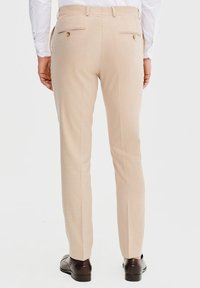 Beżowe dopasowane spodnie o gładkiej fakturze, z dwoma tylnymi kieszeniami i zapięciem na guziki, noszone z ciemnobrązowymi butami.