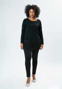 Schwarzes Langarmoberteil mit strukturiertem Muster und drapiertem Design, kombiniert mit schwarzen Leggings und silbernen Glitzer-High-Heels vor einem neutralen Hintergrund.