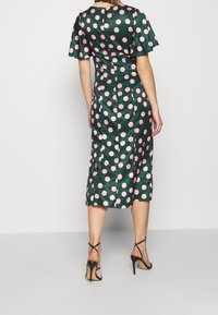 Mörkgrön klänning med korta ärmar och en figursydd midja, prydd med stora rosa prickar. Kjolen når nedanför knäet och är kombinerad med svarta högklackade skor.