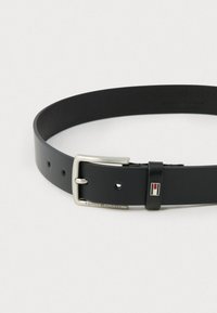 Svart lærbelte med en spenne i sølvmetall. Inkluderer et logo-accent og flere hull for størrelsejustering. Glatt tekstur gjennom hele.