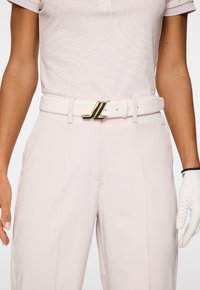 Donna che indossa pantaloni rosa pallido e una camicia coordinata, cintura bianca con fibbia dorata JL e guanto da golf bianco sulla mano destra.