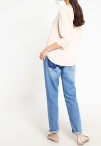 Kvinna i en ljusrosa blus, uppvikta blå jeans och blekrosa ballerinas, stående med en sidobakvy mot en vit bakgrund.