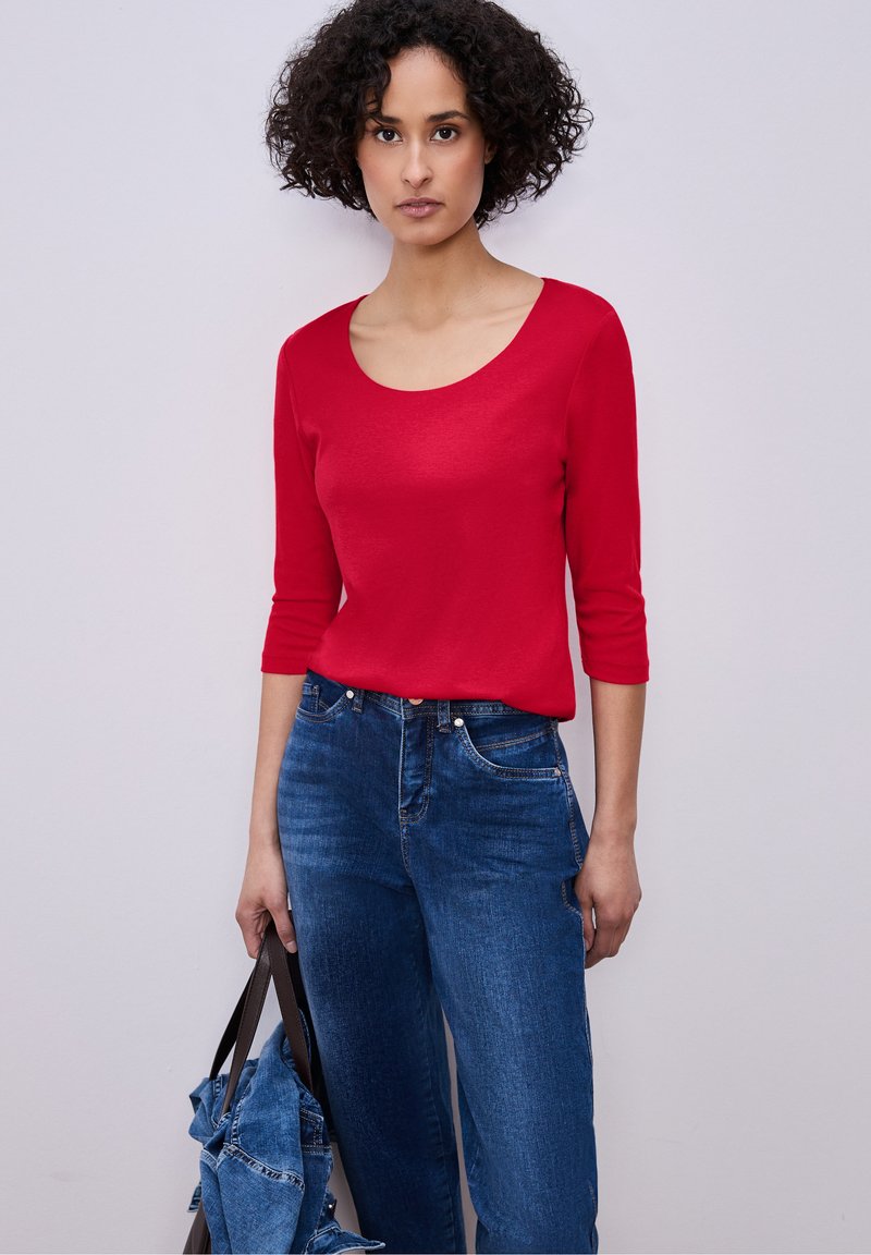 Femme aux cheveux bouclés portant une chemise rouge à manches trois-quarts et un jean bleu, tenant une veste en jean avec une sangle de sac marron.