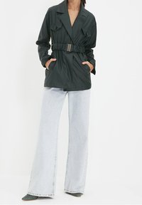 Black belted jacket with two front pockets and a notched collar, paired with light wash wide-leg jeans and green heels.