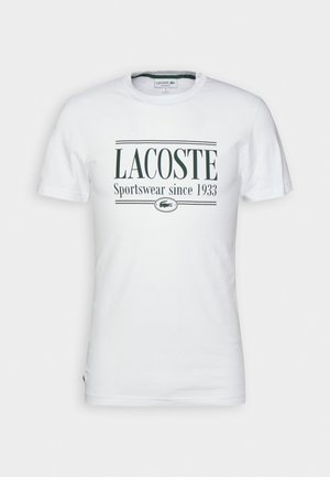 T-shirt blanc à manches courtes avec le texte « LACOSTE Sportswear depuis 1933 » et un petit logo crocodile centré sur la poitrine.