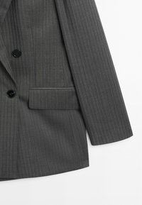 Donkergrijze krijtstreepblazer met zwarte knopen en een klepzak aan de voorkant aan de rechterkant.