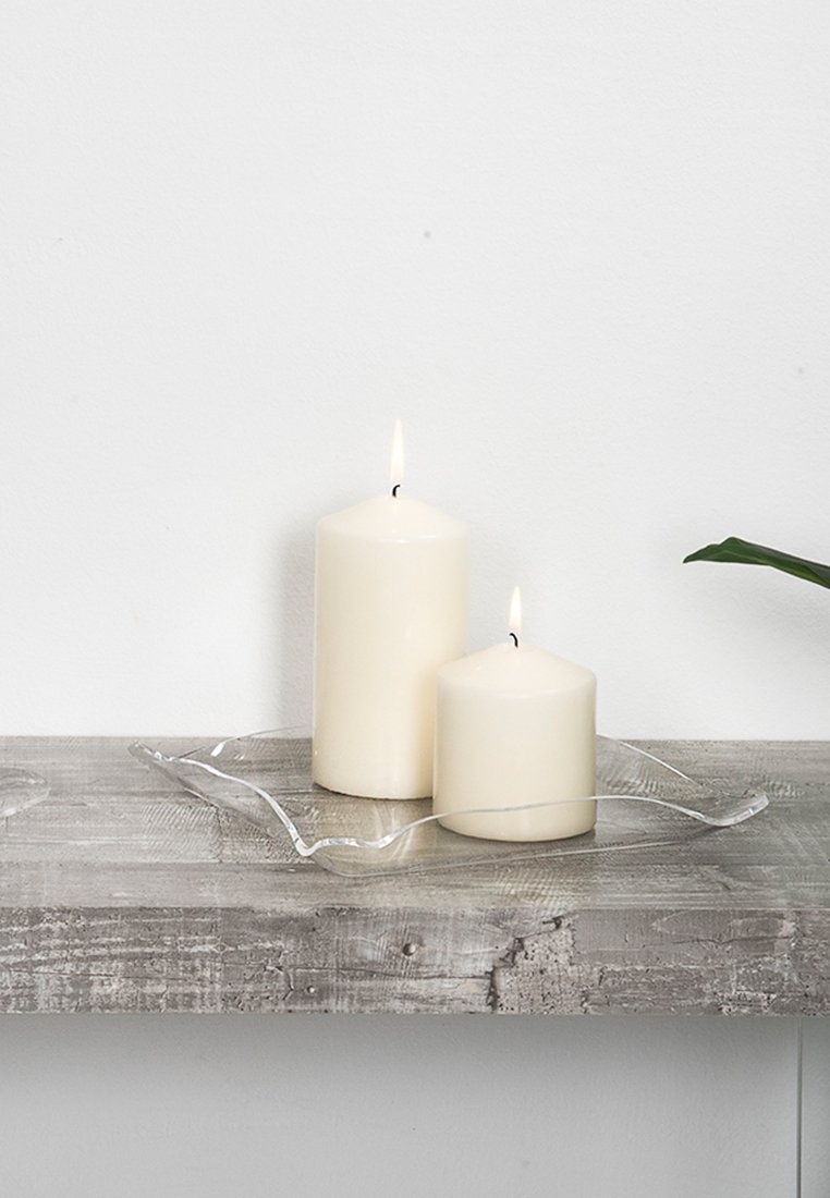 Due candele bianche, una più alta e cilindrica, l'altra più bassa e rotonda, posizionate su un vassoio di vetro trasparente a forma d'onda su una superficie in legno grigio.