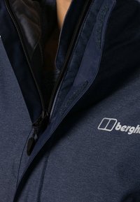 Marinblå zip-up jacka med slät yta, som har en fleecefodrad krage, kardborre-accenter och en vit logga på vänster sida.