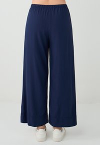 Marineblauwe wijde broek met een elastische tailleband, een gladde textuur en een relaxte pasvorm, eindigend net boven de witte sneakers aan de voeten.