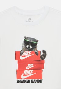 T-shirt branca com um guaxinim a usar óculos de sol verdes, sentado em caixas de sapatos Nike vermelhas empilhadas, com o texto "SNEAKER BANDIT" abaixo.