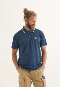 Marinblå polo med gröna detaljer, treknapps-knäppning, korta ärmar och en liten logotyp på bröstet; i bomull.