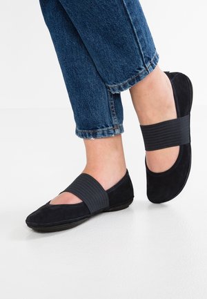 Zapatos planos negros con tiras elásticas anchas, usados con vaqueros azules remangados sobre un fondo de suelo blanco.