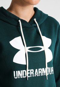 Osoba oblečená v tmavozelené mikině Under Armour s bílými šňůrkami a velkým bílým logem na hrudi.