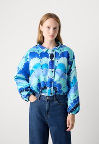 Giacca blu trapuntata con un motivo a onde in diverse tonalità, orlo elastico e fronte con bottoni e colletto. Abbinata a jeans scuri.