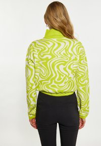Grüner und weißer gemusterter Rollkragenpullover mit einem gerippten Kragen, der ein wellenförmiges Design aufweist. Kombiniert mit schwarzen, eng anliegenden Hosen.