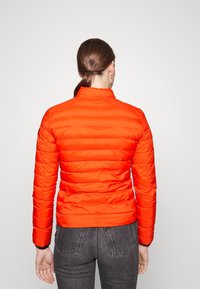 Veste légère rouge avec un col haut, texture matelassée et manches longues. Comprend des poignets élastiques et un design ajusté.