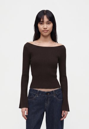 Jeune femme aux cheveux noirs et raides portant un top marron foncé à manches longues et épaules dénudées ainsi qu'un jean bleu foncé sur un fond uni.