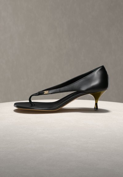 Μαύρο δερμάτινο παπούτσι με λουράκι στιλ στρινγκ και τακούνι kitten σε χρυσό χρώμα, με μικρό διακοσμητικό χρυσό λογότυπο στο λουράκι.