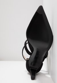 Chaussure à talon haut noire à bout pointu avec fines brides, vue du côté de la semelle contre une surface blanche et un angle de mur.