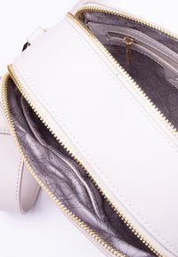 Lys beige crossbody taske lavet af tekstureret materiale, med gyldne lynlåslukninger og et mønstret interiør med flere rum.