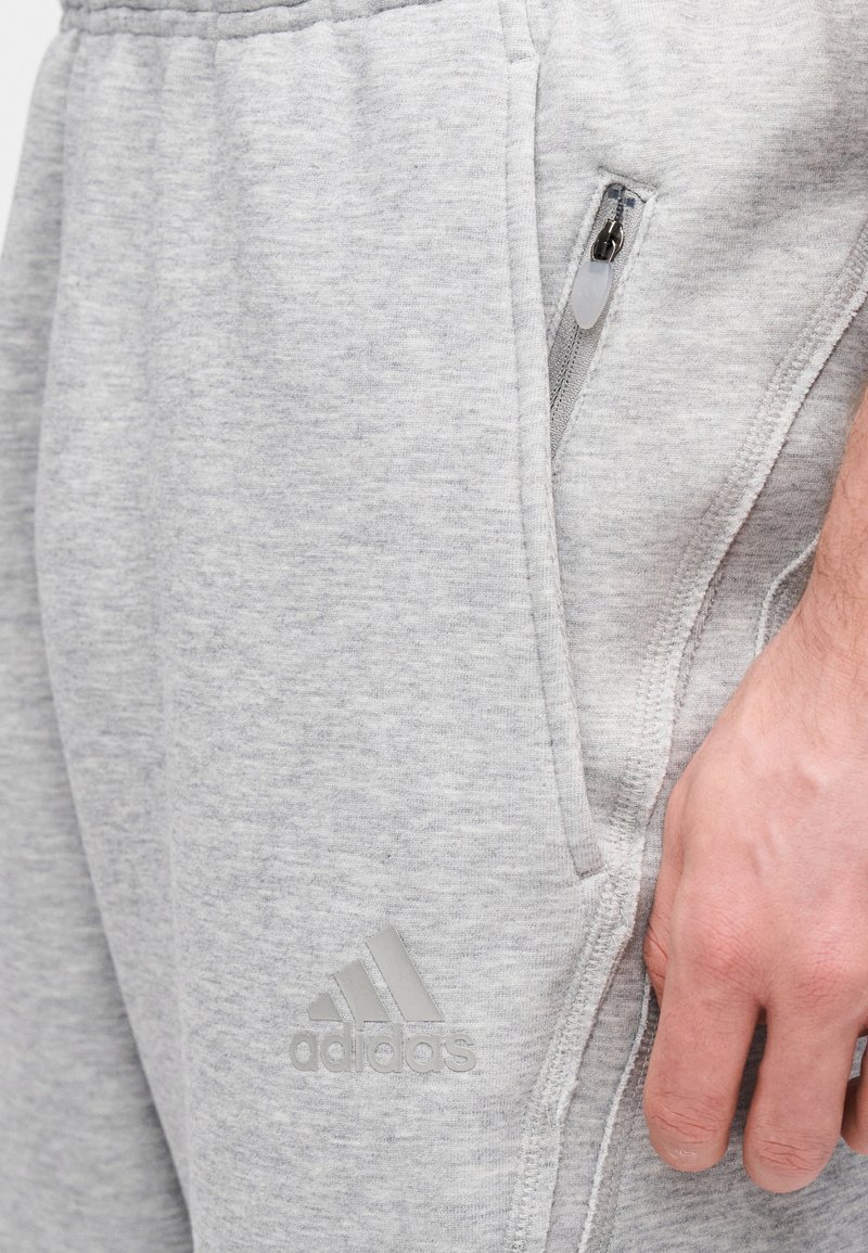 Pantalon de survêtement gris à texture lisse, coupe fuselée, poches zippées et logo Adidas réfléchissant sur la cuisse gauche.