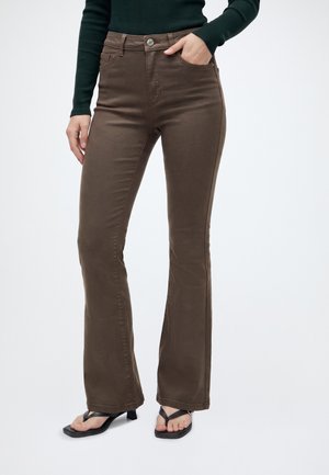 Personne portant un pantalon bootcut marron taille haute, un haut à manches longues vert foncé, et des sandales à talons noires avec des brides entre les orteils.
