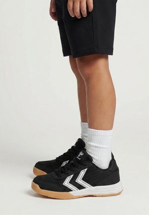 Gambe di una persona che indossa pantaloncini neri, calzini bianchi e scarpe sportive nere con logo bianco a chevron e suole in gomma.