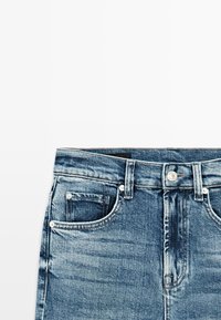 Modré džíny z denimu složené na plocho, zobrazující přední kapsy, poutka na opasek a kovové knoflíky na bílém pozadí.