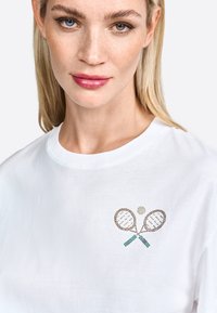 Witte katoenen t-shirt met een tennisracket graphic in Swarovski-stenen, met groene en bruine accenten, en een ronde, centrale bal detail.