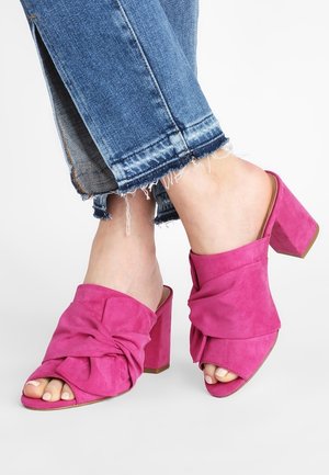 Różowe zamszowe klapki na słupku z węzłowym wzorem z przodu, w stylu odkrytych palców, z teksturowanym wykończeniem. Widoczne dżinsy z postrzępionymi nogawkami.
