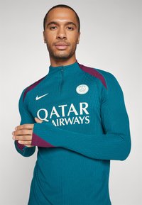 Teal lange mouwen trui met hoge kraag, voorzien van paarse accenten, een wit logo en de tekst "QATAR AIRWAYS" op de voorkant. Gestructureerde stof.