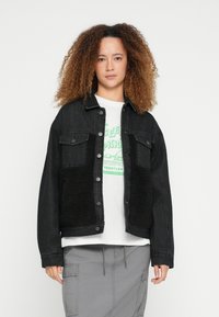 Fekete farmerrövid kabát fekete bolyhos panelekből, patentos gombokkal, mellzsebekkel és laza fazonban, fehér grafikás póló fölött viselve.