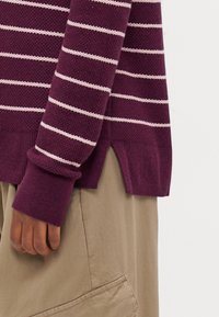 Lila randig tröja med vita horisontella ränder, texturerad tyg, ribbade muddar och sidofickor, i kombination med beige byxor som har en ficka.