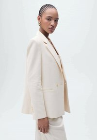 Beige přizpůsobený blazer s jedním knoflíkem, klopovými límečky a bočními kapsami. Hladká textura, v kombinaci s odpovídajícími kalhotami.