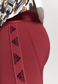 Burgundowe legginsy wykonane z elastycznego materiału o gładkiej strukturze. Posiadają czarne logo Adidas po bokach. Zbliżenie.