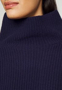 Pull à col roulé bleu marine foncé, texture côtelée, matière douce, col haut, coupe ajustée avec de subtiles lignes verticales.