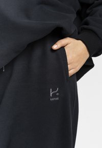 Hand in der Tasche eines schwarzen Sweatshirts, auf dem das gestickte "kamak"-Logo auf dem Stoff vor einem schlichten Hintergrund zu sehen ist.