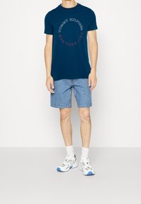 Námořnická modrá tričko s krátkým rukávem a kruhovým logem s textem "TOMMY HILFIGER NEW YORK CITY." V kombinaci s světle modrými džínovými kraťasy a bílými teniskami.