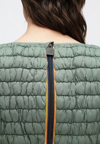 Gewatteerd groen textieljack met een zichtbare rits en oranje, gele en marineblauwe strepen langs de rugnaad. Gestructureerd oppervlak met pofjes.