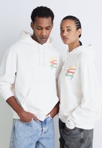 Sudaderas blancas con bolsillos frontales presentan texto bordado colorido en rojo, amarillo y verde. Ambos llevan jeans azul claro y gris.