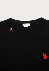 Fekete V nyakú pulóver, piros hímezett póló játékos logóval a mellen és kis piros, fehér, kék csík részlettel a nyak környékén.