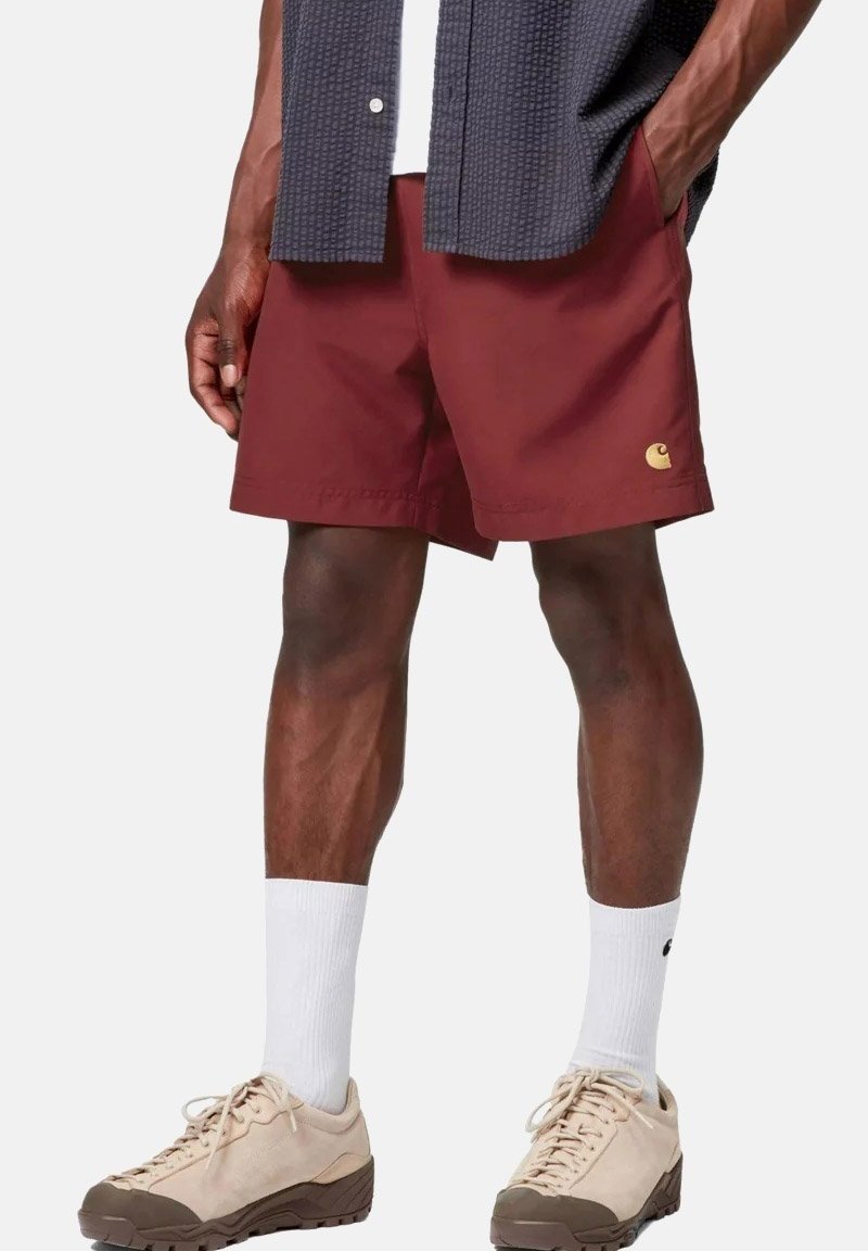 Mann trägt bordeauxrote Shorts, weiße Socken, beige Sneakers und ein dunkles strukturiertes Hemd mit den Händen in den Taschen.