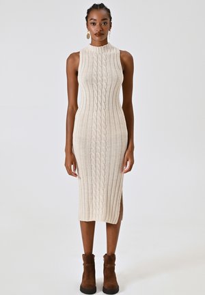 Abito in maglia beige a coste con colletto alto, caratterizzato da motivi a treccia, spacchi laterali e una vestibilità aderente. Stivali alla caviglia marroni completano il look.