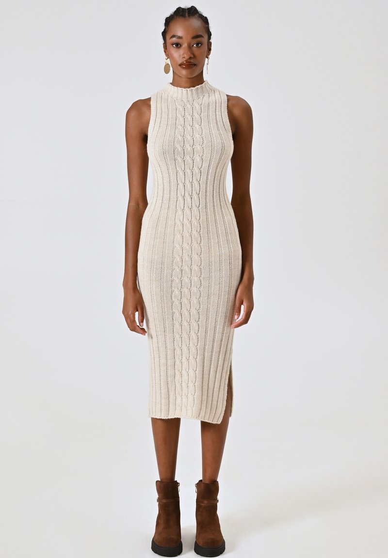 Abito in maglia beige a coste con colletto alto, caratterizzato da motivi a treccia, spacchi laterali e una vestibilità aderente. Stivali alla caviglia marroni completano il look.