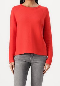 Pull en maille rouge côtelé à manches longues, col rond et coupe décontractée, associé à un jean en denim gris.