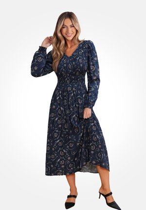 Vestido azul marino con estampado floral, cuerpo fruncido, mangas largas y falda fluida. Llevado con mulas negras de tacón, con punta afilada.
