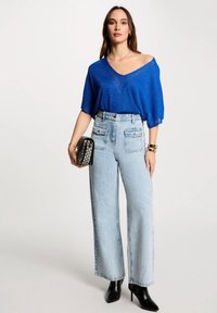 Elektrisch blaue Kurzarm-Strickbluse mit V-Ausschnitt, kombiniert mit hellen, hochgeschnittenen, weiten Jeans und schwarzen spitzen Stiefeletten.