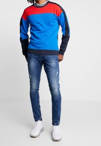 Blue, red, and navy sweatshirt with a round neck and stripes, paired with distressed light blue jeans and white sneakers.